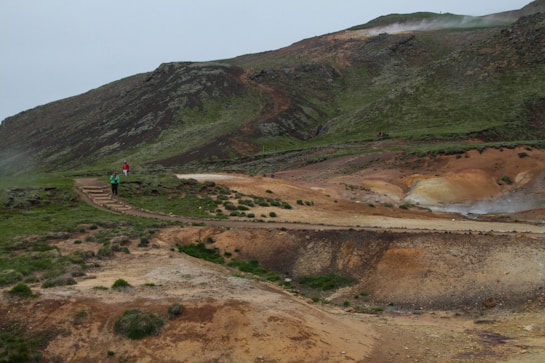 A rugged, mountainous landscape with green and brown hues features a hiking trail winding through it. Small groups of people walking along the path can be seen, indicating a sense of exploration and adventure. The terrain appears natural and slightly barren, with subtle steam rising in the background.