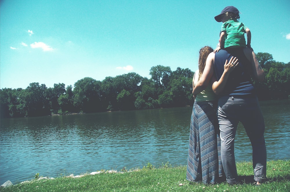 father, mother, and son standing on grass lawn near at body of water during daytime