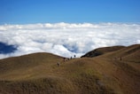 A group of young trekkers pausing on a lush green mountain ridge under a bright blue sky.