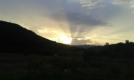 A landscape capturing the sun setting behind a range of low hills, casting dramatic rays across the sky. Dark silhouettes of hills and some trees are visible, contrasted against the warm glow of the sunset. The sky features a mix of clouds, adding texture and depth to the scene.