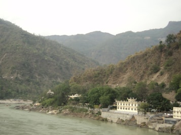 A river flows alongside a mountainous landscape, with hills covered in green vegetation. Several buildings, including a temple, are situated near the riverbank, surrounded by trees.