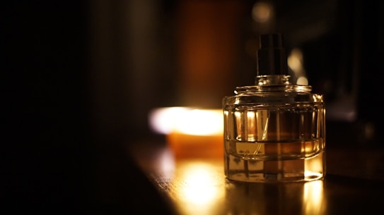 A glass perfume bottle is illuminated by warm, soft lighting, reflecting on a wooden surface. A blurred background adds depth, with a subtle play of light and shadow.