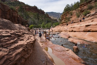 People are enjoying a natural waterway surrounded by red rock formations and cliffs in a picturesque canyon setting. Some are swimming or wading in the water, while others walk along the rock paths. The area is lush with green trees and the sky is bright, indicating a sunny day.