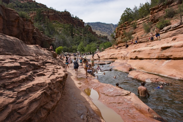 People are enjoying a natural waterway surrounded by red rock formations and cliffs in a picturesque canyon setting. Some are swimming or wading in the water, while others walk along the rock paths. The area is lush with green trees and the sky is bright, indicating a sunny day.