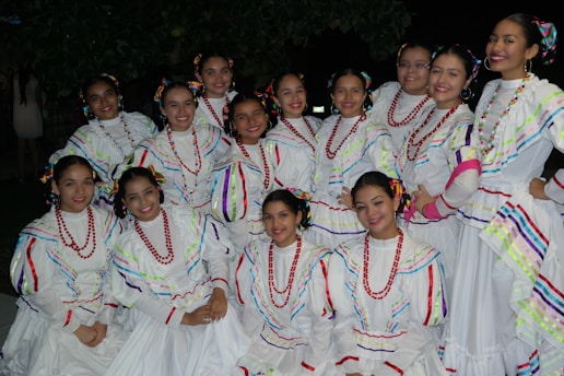 A group of women dressed in traditional white dresses with colorful stripes and red bead necklaces, arranged in a semi-circle formation. They are smiling and appear to be posing for the photo, taken outdoors at night with trees in the background.