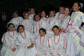 A group of women dressed in traditional white dresses with colorful stripes and red bead necklaces, arranged in a semi-circle formation. They are smiling and appear to be posing for the photo, taken outdoors at night with trees in the background.