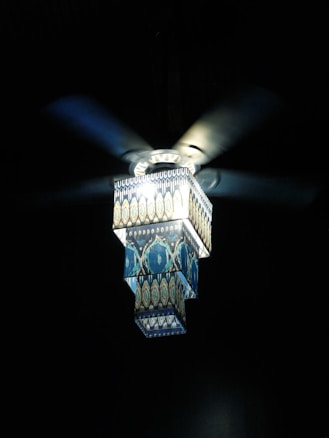 A decorative ceiling fan with a geometric, patterned light cover is illuminated against a dark background. The light has a layered design with intricate blue and gold patterns, creating a visually striking contrast with the surrounding darkness.