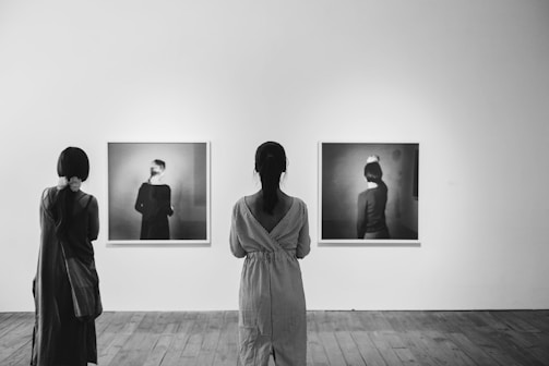 In a gallery, two people are observing black and white photographs displayed on a white wall. The room features wooden flooring, and the atmosphere is quiet and contemplative.