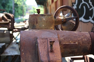 An old, rusted industrial machine with visible wear and corrosion. The machinery appears to be outdoors, with blurred greenery and industrial structures in the background. The image captures the weathered texture and intricate details of the metalwork, including bolts and a large wheel.