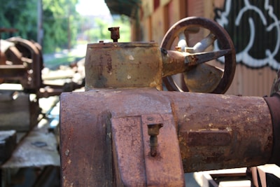 An old, rusted industrial machine with visible wear and corrosion. The machinery appears to be outdoors, with blurred greenery and industrial structures in the background. The image captures the weathered texture and intricate details of the metalwork, including bolts and a large wheel.