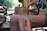 An old, rusted industrial machine with visible wear and corrosion. The machinery appears to be outdoors, with blurred greenery and industrial structures in the background. The image captures the weathered texture and intricate details of the metalwork, including bolts and a large wheel.
