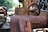 An old, rusted industrial machine with visible wear and corrosion. The machinery appears to be outdoors, with blurred greenery and industrial structures in the background. The image captures the weathered texture and intricate details of the metalwork, including bolts and a large wheel.