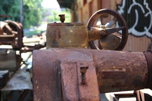 An old, rusted industrial machine with visible wear and corrosion. The machinery appears to be outdoors, with blurred greenery and industrial structures in the background. The image captures the weathered texture and intricate details of the metalwork, including bolts and a large wheel.