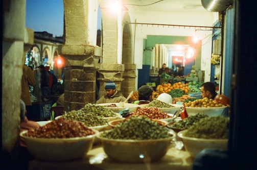 A busy market scene with several large bowls filled with olives and other produce. There are multiple vendors, some seated, engaging in business. The lighting suggests it is either early morning or late evening with artificial lights illuminating the area, casting a warm glow. Stone columns and arched pathways enhance the rustic and traditional atmosphere.