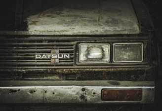 A worn-out front view of a vintage Datsun car, showcasing rust and dirt on its metal surface. The headlights appear dull and aged, with a faded emblem at the center of the grille. The paint is weathered, with visible patches of corrosion.
