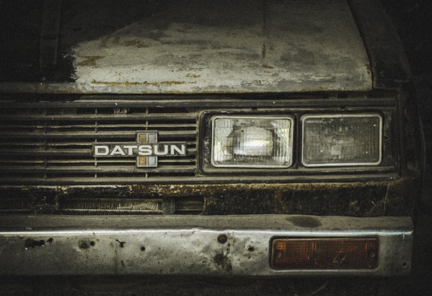 A worn-out front view of a vintage Datsun car, showcasing rust and dirt on its metal surface. The headlights appear dull and aged, with a faded emblem at the center of the grille. The paint is weathered, with visible patches of corrosion.