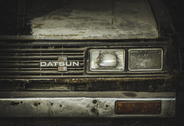A worn-out front view of a vintage Datsun car, showcasing rust and dirt on its metal surface. The headlights appear dull and aged, with a faded emblem at the center of the grille. The paint is weathered, with visible patches of corrosion.