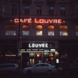 A night-time street scene featuring the facade of Café Louvre, illuminated by neon lights. The building's first floor windows reflect a warm glow from inside. Below the neon sign, there are several parked cars and an entrance to the cafe and restaurant area.