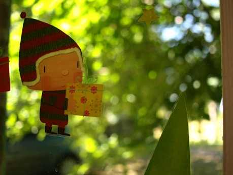 A cute cartoon character is holding a yellow gift box with floral patterns. The character is wearing a red and green striped winter hat with ear flaps. In the background, there's a blurred view of lush green foliage and a dimly lit scene, suggesting a garden or natural setting.