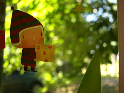 A cute cartoon character is holding a yellow gift box with floral patterns. The character is wearing a red and green striped winter hat with ear flaps. In the background, there's a blurred view of lush green foliage and a dimly lit scene, suggesting a garden or natural setting.
