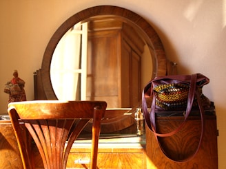 A wooden vanity table is adorned with a round mirror and a colorful woven bag. A warm light bathes the scene, casting soft shadows on the wall. A small, decorative figurine is placed on the left side of the table.