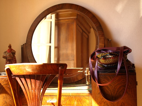 A wooden vanity table is adorned with a round mirror and a colorful woven bag. A warm light bathes the scene, casting soft shadows on the wall. A small, decorative figurine is placed on the left side of the table.