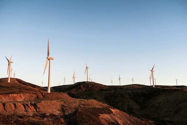 Wind turbines are scattered across a hilly, arid landscape under a clear blue sky. The turbines are tall and white, and their blades stand out against the setting sun, casting long shadows. The terrain is rugged with visible dirt paths and rocky outcrops.