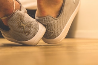 A close-up view of gray sports shoes with a woven texture, featuring a silhouette logo on the heel. The shoes are positioned on a light wooden floor, with a blurred background adding depth to the image.