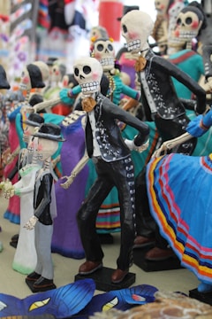 A collection of colorful Day of the Dead figurines, crafted to resemble skeletons in festive attire. The figures are adorned with bright clothing and accessories, featuring detailed skeletal faces with prominent eye sockets and teeth, conveying a sense of celebration and tradition.