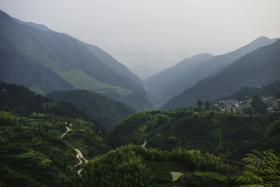 The image depicts a mountainous landscape with a winding road cutting through it, surrounded by lush green trees and vegetation. The mountains rise majestically on either side of the road, creating a sense of depth and scale. In the distance, beyond the verdant hills, there is a small town nestled among the mountains.