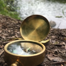 A brass compass lying on a forest floor near the edge of a body of water. The compass is open, showing the directional markings clearly. The surroundings include dry leaves, twigs, and a blurred background of water and greenery.