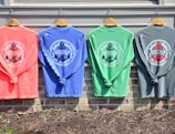 Four colorful long-sleeve shirts hang on wooden hangers against a light-colored wall. Each shirt features a nautical-themed design with the words 'St. Michaels Maryland' and an anchor emblem in the center. The shirts are in coral, blue, green, and gray tones, and are displayed above a brick wall with some mulch and soil visible below.