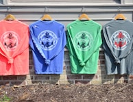 Four colorful long-sleeve shirts hang on wooden hangers against a light-colored wall. Each shirt features a nautical-themed design with the words 'St. Michaels Maryland' and an anchor emblem in the center. The shirts are in coral, blue, green, and gray tones, and are displayed above a brick wall with some mulch and soil visible below.