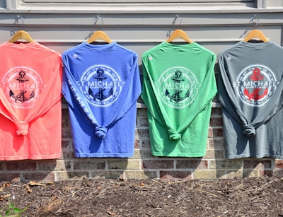 Four colorful long-sleeve shirts hang on wooden hangers against a light-colored wall. Each shirt features a nautical-themed design with the words 'St. Michaels Maryland' and an anchor emblem in the center. The shirts are in coral, blue, green, and gray tones, and are displayed above a brick wall with some mulch and soil visible below.