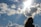 A silhouette of a person with flowing hair is positioned against a bright sky filled with scattered clouds. The sun beams through the clouds, casting a radiant light, while a bird soars in the distance.