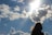 A silhouette of a person with flowing hair is positioned against a bright sky filled with scattered clouds. The sun beams through the clouds, casting a radiant light, while a bird soars in the distance.