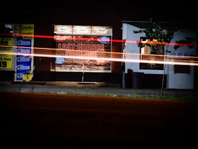A night scene featuring a convenience store or package store with bright signage displaying 'Last Chance Package' alongside advertisements for various beer brands. Long exposure light trails, possibly from passing vehicles, create streaks across the image. A small tree is visible near the building, with a vending machine or display on the right side of the entrance.