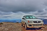 A Suzuki car is parked on a rugged gravel road, surrounded by mountain peaks under a cloudy sky. The car's white exterior is slightly dirty, likely due to the off-road conditions.