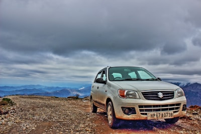 A Suzuki car is parked on a rugged gravel road, surrounded by mountain peaks under a cloudy sky. The car's white exterior is slightly dirty, likely due to the off-road conditions.