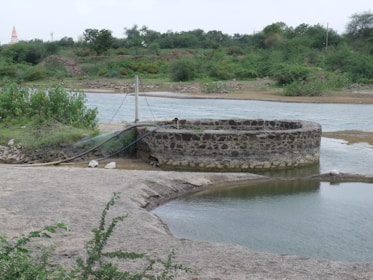 A circular stone well is situated near a body of water in a rural landscape. The surrounding area is lush with green vegetation and bushes. There are pipes connected to the well, likely for water drawing purposes. In the background, trees and greenery dominate the scene, providing a natural setting.