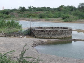 A circular stone well is situated near a body of water in a rural landscape. The surrounding area is lush with green vegetation and bushes. There are pipes connected to the well, likely for water drawing purposes. In the background, trees and greenery dominate the scene, providing a natural setting.