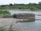A circular stone well is situated near a body of water in a rural landscape. The surrounding area is lush with green vegetation and bushes. There are pipes connected to the well, likely for water drawing purposes. In the background, trees and greenery dominate the scene, providing a natural setting.