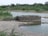 A circular stone well is situated near a body of water in a rural landscape. The surrounding area is lush with green vegetation and bushes. There are pipes connected to the well, likely for water drawing purposes. In the background, trees and greenery dominate the scene, providing a natural setting.
