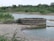 A circular stone well is situated near a body of water in a rural landscape. The surrounding area is lush with green vegetation and bushes. There are pipes connected to the well, likely for water drawing purposes. In the background, trees and greenery dominate the scene, providing a natural setting.
