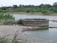 A circular stone well is situated near a body of water in a rural landscape. The surrounding area is lush with green vegetation and bushes. There are pipes connected to the well, likely for water drawing purposes. In the background, trees and greenery dominate the scene, providing a natural setting.