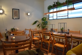 A cozy cafe interior with wooden furniture and a warm ambiance. Several wooden chairs and tables are neatly arranged along with small decorative plants. A framed picture hangs on the wall, surrounded by small wall-mounted lights. The window is adorned with hanging plants, adding a touch of greenery to the space.