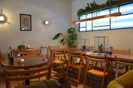 A cozy cafe interior with wooden furniture and a warm ambiance. Several wooden chairs and tables are neatly arranged along with small decorative plants. A framed picture hangs on the wall, surrounded by small wall-mounted lights. The window is adorned with hanging plants, adding a touch of greenery to the space.