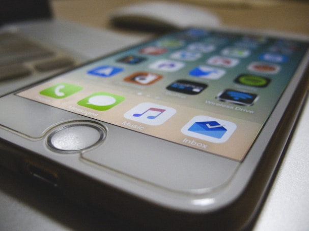 A close-up view of a smartphone displaying various colorful app icons on its screen. The focus is on the lower portion of the phone, highlighting the home button and a row of apps including Messages, Music, and Inbox.