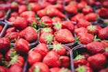 A collection of ripe, red strawberries fills clear plastic containers. The strawberries are vibrant in color with their seeds and green leaves clearly visible. The arrangement is densely packed, showing an abundance of fruit.
