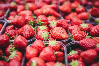 A collection of ripe, red strawberries fills clear plastic containers. The strawberries are vibrant in color with their seeds and green leaves clearly visible. The arrangement is densely packed, showing an abundance of fruit.